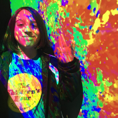 A child wearing a Children's Hour tshirt, standing in a colorful projection of light.