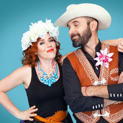 Alisha Gaddis and Lucky Diaz, posed in white hats. Lucky is looking at Alisha. There is a blue background.