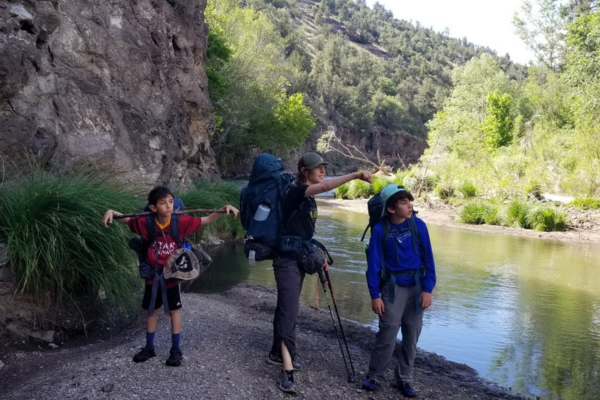 On the bank of the Gila River, a mom is pointing to the right and her two young sons are on either side of her looking in the direction she is pointing. They are on the gravel bank against a cliff with the Gila River on the right side of them, twisting away into the distance up the frame. 