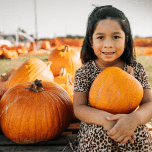 A girl with dark brown hair, a leopard print dress, and a big smile is holding a pumpkin with many pumpkins next to her.