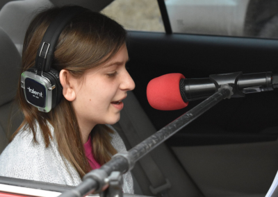 A child smiling and singing into a microphone while sitting in the back of her car. Child is wearing headphones and we see her from the side.