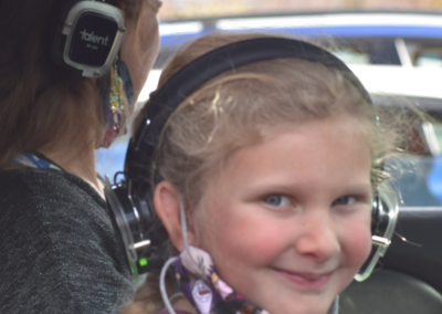 A child smiling while sitting in the front passenger seat of a car. Child is wearing headphones and we see her from the side looking directly at the camera.