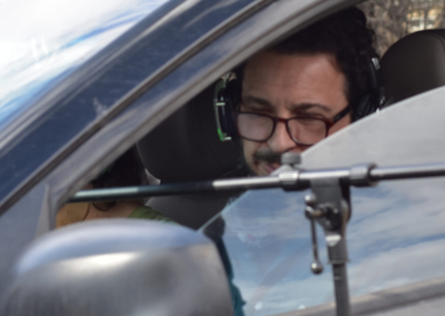 A man wearing headphones sitting in the driver's seat of a car, with a microphone stand coming through the window. The man is concentrating.