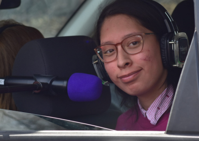 A child smiling into a microphone while sitting in the back of her car. Child is wearing headphones and we see her from the front.