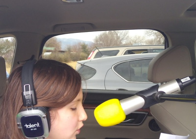 A child singing into a microphone while sitting in the back of her car. Child is wearing headphones and holding a script.