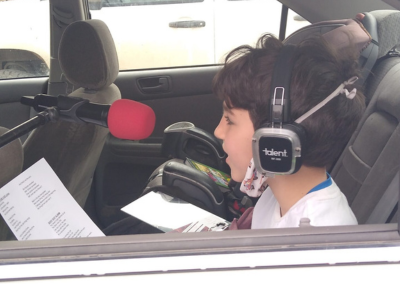 A child singing into a microphone while sitting in the back of her car. Child is wearing headphones and we see him from the side through an open car window.