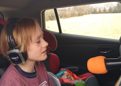 A child singing into a microphone while sitting in the back of her car. Child is wearing headphones and we see him from the front.