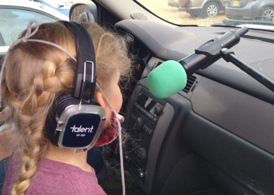 A child singing into a microphone while sitting in the back of her car. Child is wearing headphones and we see her from behind.