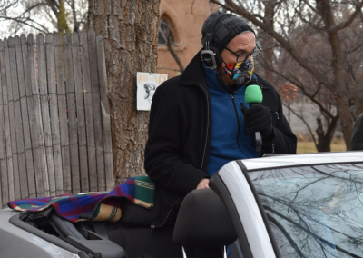 The director sitting on the back of his convertible car, bundled up because it is cold and cloudy, with a microphone in front of his mouth and wearing a mask.