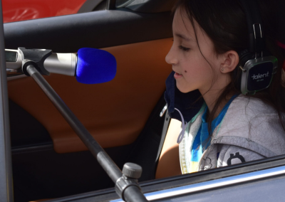 A child sitting in front of a microphone while sitting in the back of her car. Child is wearing headphones and we see her from the side.