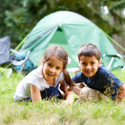 Camping With Kids - Cover Two children with brown hair, about 5 years old, a girl and a boy sitting on grass in front of a green tent.