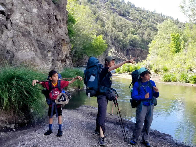 Camping with Kids - Isaac in Gila Isaac and his brother stand with their mom at the edge of the Gila River, all are wearing backpacking backpacks, and the mom is pointing to something in the distance to the right, and Isaac looking where she is pointing.