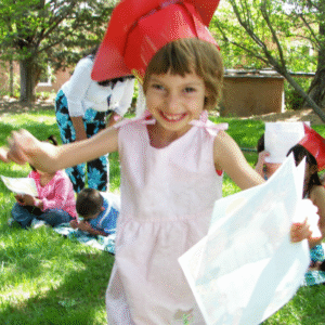 A 5 year old child wearing a red graduation cap, pink dress, holding a paper diploma, and running toward camera smiling. Child is a little blurry.