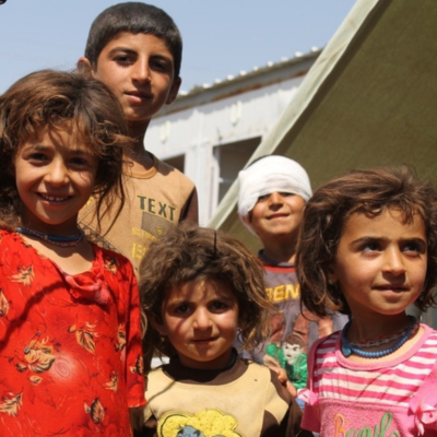 Five children in a refugee camp. One in back has bandaged eyes. Kids are smiling but it's clear life is difficult.