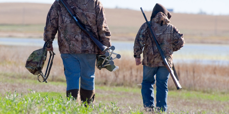 A hunting child walking with an adult away from the camera, wearing camo and carrying guns. 
